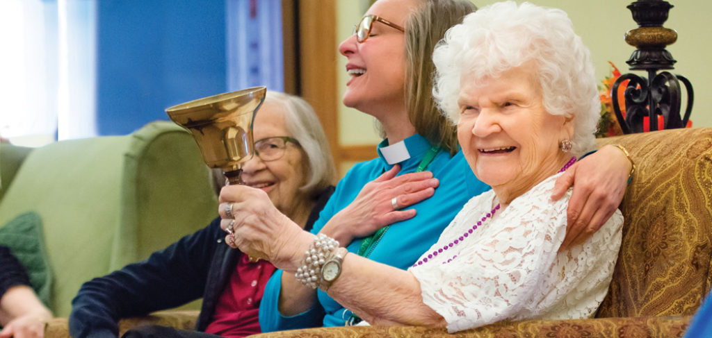 Seniors and chaplain play a handbell while laughing and smiling