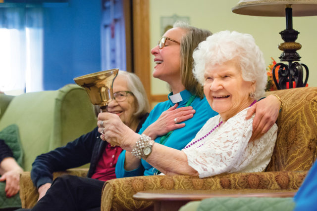 Seniors and chaplain play a handbell while laughing and smiling