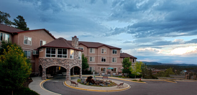 Exterior view of Elk Run Assisted Living in Evergreen, CO, featuring a stone archway, a circular driveway with a fountain, and scenic mountain surroundings under a cloudy sky